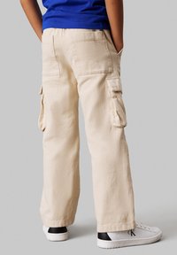 Pantalons cargo beiges avec poches latérales et poches arrière à patte. Fabriqués en tissu texturé, avec une coupe droite et une coupe décontractée. Associés à des chaussures blanches.