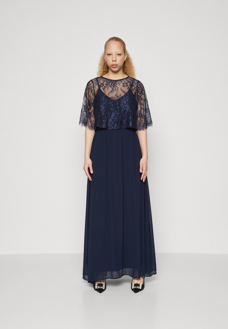 Μάξι φόρεμα ναυτικού μπλε με δαντελένια επικάλυψη, κοντά φουσκωτά μανίκια και απαλό κάτω μέρος από chiffon. Τα διακοσμημένα μαύρα μυτερά παπούτσια ολοκληρώνουν την εμφάνιση.