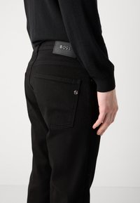 Crne traperice s uskim krojem, s vidljivom etiketom na struku, stražnjim džepovima i suptilnim detaljima šivanja.