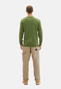 Grön tröja med långa ärmar, rundad hals och ett detaljerat mönster på ryggen, kombinerad med beige cargobyxor och gula kängor. Enkel och funktionell design.