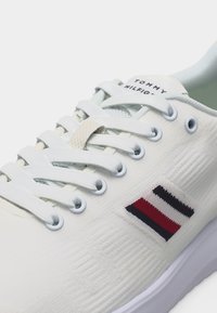 Κοντινή λήψη λευκού αθλητικού παπουτσιού με λευκά κορδόνια, λογότυπο Tommy Hilfiger στη γλώσσα και λεπτομέρεια με κόκκινη, λευκή και ναυτική σημαία στην πλαϊνή πλευρά.