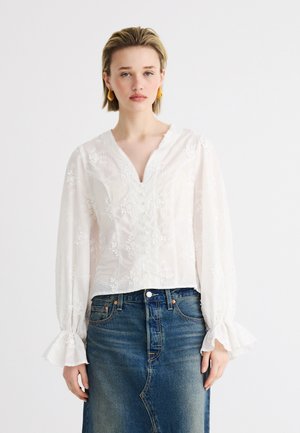 Jeune femme portant une blouse blanche brodée à manches longues avec des poignets à volants et une jupe en jean bleue, debout devant un fond blanc uni.