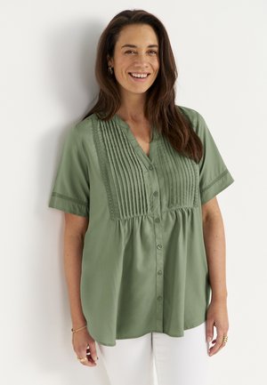 Uśmiechnięta kobieta z długimi brązowymi włosami, ubrana w zieloną, krótką, zapinaną na guziki bluzkę i białe spodnie, stojąca na tle białej ściany.