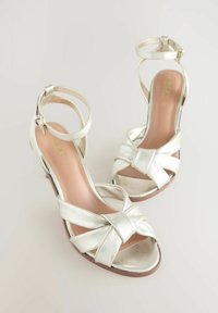 Zilveren metallic hoge hakken sandalen met een gedraaid knoopontwerp aan de voorkant, een verstelbare enkelband en een gladde lederen afwerking.