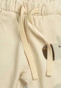 Pantalon de survêtement beige clair en tissu texturé, taille à cordon avec liens plats tissés et un discret motif animalier en brun foncé.