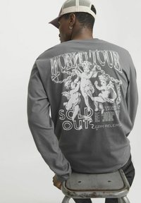 Langärmliges graues Shirt mit Grafikdruck auf dem Rücken, das Putten und den Text "WORLD TOUR SOLD OUT" in kontrastierendem Weiß zeigt.