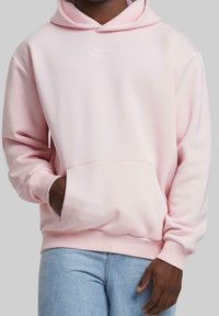 Man i en ljusrosa hoodie med en framficka och ljusblå jeans, hand vilande inuti fickan mot en enkel bakgrund.