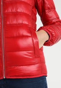Giacca a vento rossa con superficie liscia e lucida, cuciture orizzontali e tasca laterale. I polsini sono elasticizzati per una vestibilità aderente.