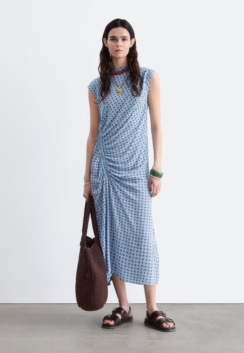 Femme portant une robe midi bleue à motifs sans manches, tenant un grand sac tricoté marron, portant des sandales noires et des colliers superposés.