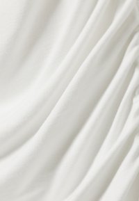 Mykt hvitt stoff med subtile folder og en mild tekstur som lager glatte, flytende linjer over flaten.