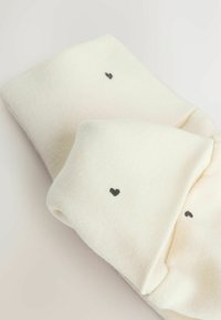 Cremefarvede sokker lavet af blødt stof, med små sorte hjerte-motiver på ydersiderne. Glat tekstur med rullede manchetter.