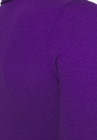 Purple knitted garment shows a close-up of the fabric texture, featuring a smooth, even knit pattern with a slightly ribbed neckline.