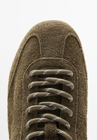 Suede sneaker in olive green with textured surface. Features grey laces with white accents and a rounded toe cap design.