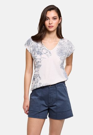 Mujer con camiseta blanca de escote en V con estampado botánico azul y pantalones cortos de talle alto azul marino, de pie contra un fondo liso.