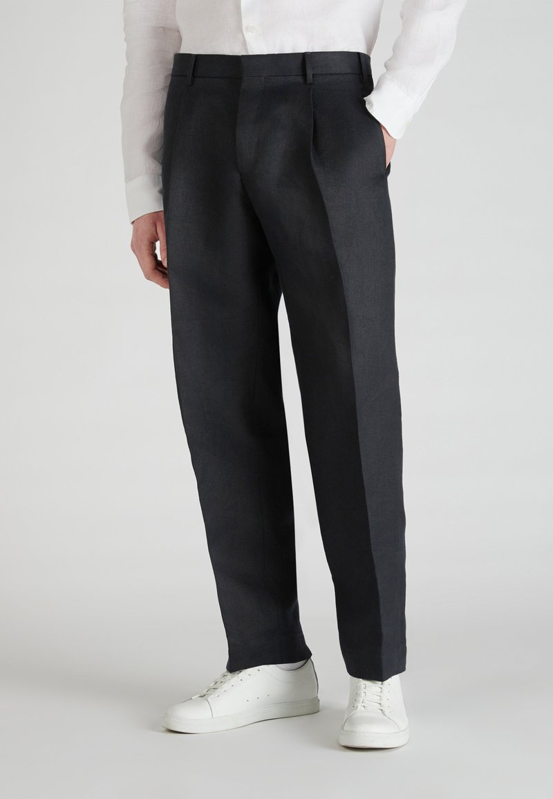 Pantalones de lino gris oscuro con un corte holgado, cuentan con dos pliegues frontales y bolsillos laterales, combinados con zapatillas blancas.