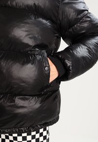 Černá péřová bunda s lesklým povrchem, disponující kapsou na zip, žebrovanými manžetami a přiléhavým střihem v pase.