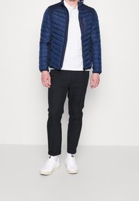 Daunenjacke in Dunkelblau mit Steppmuster, Reißverschluss vorne und Seitentaschen. Getragen über einem weißen Polo mit Muster und schwarzen, zulaufenden Hosen. Weiße Sneakers.