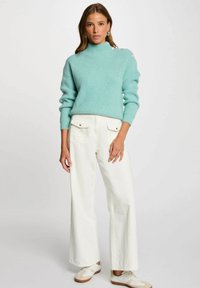 Maglione turchese lavorato a maglia con collo alto e bottoni decorativi sulla spalla, abbinato a pantaloni bianchi a gamba larga e sneakers color crema.