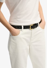 Cinturón de cuero negro con una hebilla rectangular dorada, llevado alrededor de la cintura, combinado con pantalones de lienzo blancos y una camiseta blanca sencilla.