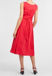 Robe sans manches rouge avec un détail de taille nouée, silhouette en A, fabriquée en tissu lisse, tombe sous le genou, associée à des talons carrés noirs.