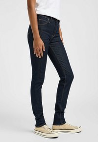 Jeans skinny de mezclilla oscura con detalles de costura naranja. Cuenta con cinco bolsillos y una cintura de altura media. Combinados con zapatillas beige.