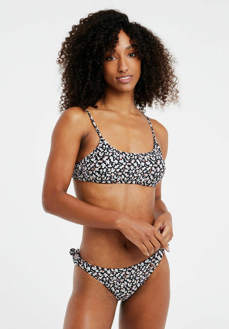 Bikini nero con motivo floreale bianco e rosa; top in stile bralette con spalline regolabili, culotte a vita bassa abbinate con dettaglio dei lacci laterali.