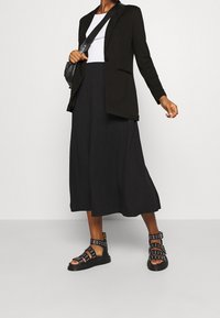 Blazer noir sur un haut blanc, associé à une jupe midi noire fluide et des sandales noires à lanières avec des accents métalliques.