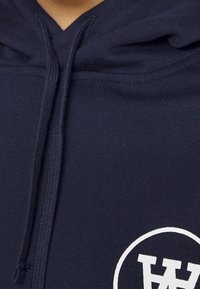 Mörkblå hoodie med känguruficka, ribbade ärmslut och en vit cirkulär logotyp på bröstet. Tillverkad av en strukturerad bomullsblandning.