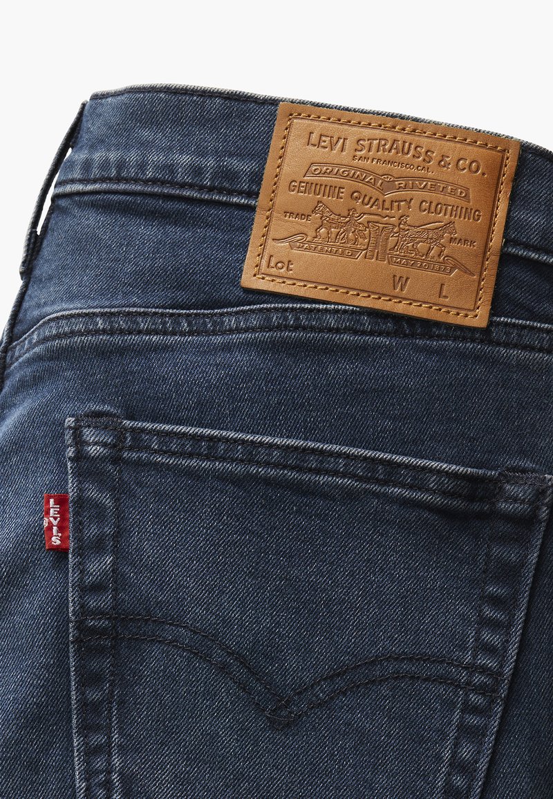 Jean en denim foncé avec une étiquette en cuir tan portant la mention "LEVI STRAUSS & CO." avec des graphiques en relief, tab rouge emblématique sur la poche arrière.