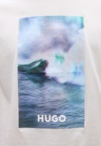 Θολό αφηρημένο κύμα ωκεανού σε αποχρώσεις του μπλε και του πράσινου με τη λέξη "HUGO" σε λευκό στο κάτω μέρος  πάνω σε λευκό φόντο υφάσματος.