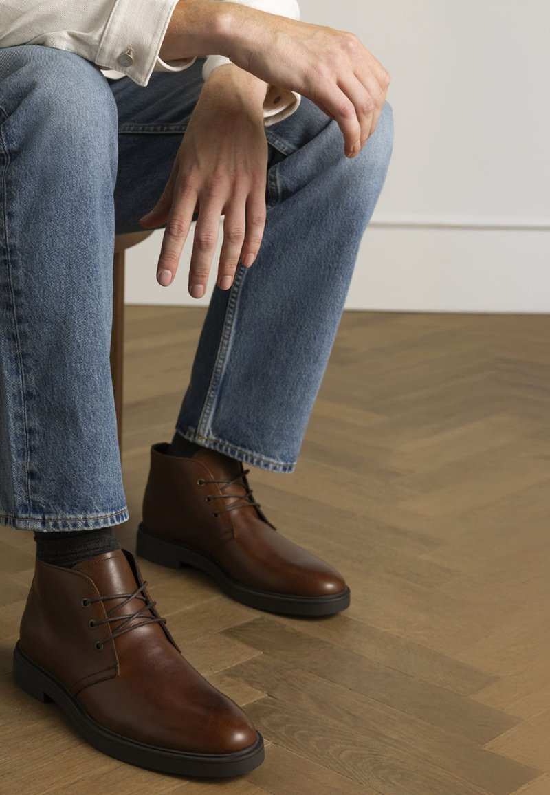 Braune Lederstiefeletten mit runden Zehen, Schnürung und strukturiertem Finish. Tragen zu blauen Jeans, auf einem Holzboden.