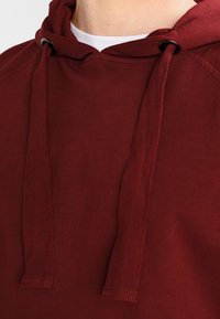 Burgundská mikina s jemnou textúrou, s šnúrkami pri krku a decentným rebrovaním na manžetách a lemovaní.