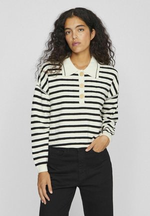 Suéter a rayas en crema y negro, con un cuello clásico, cuatro botones y puños acanalados. Combinado con pantalones negros.