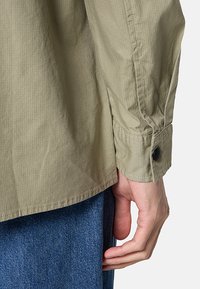 Leichtes olivgrünes Hemd mit Knopfleiste und gerippter Textur, ausgestattet mit einem strukturierten Kragen und geknöpften Manschetten. Über blauer Jeans getragen.