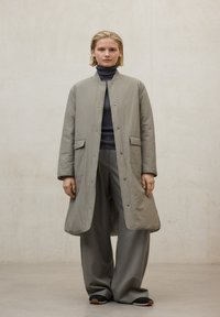 Abrigo largo de color gris claro acolchado con cuello redondo, cierre de botones y bolsillos laterales, combinado con pantalones grises sueltos a juego y zapatos negros.