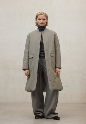 Abrigo largo de color gris claro acolchado con cuello redondo, cierre de botones y bolsillos laterales, combinado con pantalones grises sueltos a juego y zapatos negros.