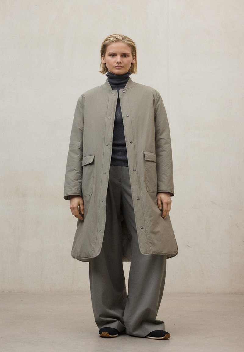 Abrigo largo de color gris claro acolchado con cuello redondo, cierre de botones y bolsillos laterales, combinado con pantalones grises sueltos a juego y zapatos negros.