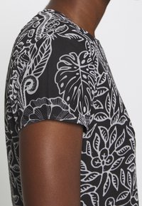 Gros plan d’une personne portant une chemise noire avec des motifs floraux et feuillages blancs détaillés sur la manche et l’épaule.