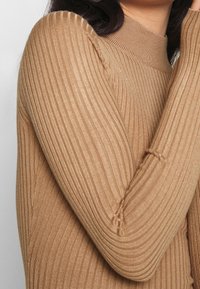 Mujer con una blusa ajustada de canal�é en color camel, de manga larga y cuello alto, con el brazo doblado cubriendo parte de su cara.