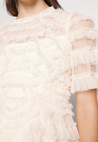 Blouse crème transparente avec tissu en tulle à volants superposés et manches courtes bouffantes sur la partie supérieure du torse d’une femme.