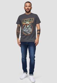 Camiseta gris con un gráfico de "El Imperio Contraataca" que presenta un diseño de Boba Fett. Combinada con jeans azul oscuro y zapatillas blancas.