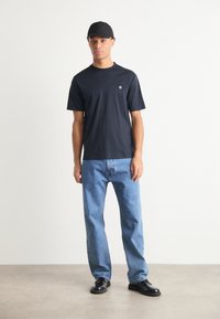 T-shirt noir à manches courtes avec un petit logo sur la poitrine, associé à un jean droit bleu clair et des chaussures noires.