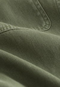 Gros plan sur un tissu vert olive avec des coutures visibles et un tissage texturé, montrant des plis et un détail de couture.