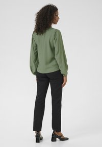 Olijfgroene blouse met lange mouwen en geplooide schouders, gecombineerd met zwarte slim-fit broek en zwarte hakken, gezien van achteren.
