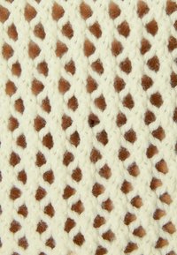 Stickad tyg i krämfärgad nyans med ett diamantformat öppet vävningsmönster som avslöjar en texturerad brun yta under. Mjuk, fiberrik textur.