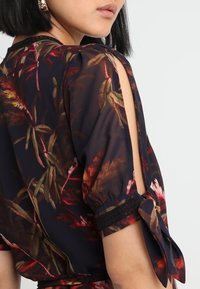 Blůza s květinovým vzorem z tmavé tkaniny, s vyříznutými rameny, černými krajkovými detaily a zavázanými rukávy. Jemná, splývavá textura.