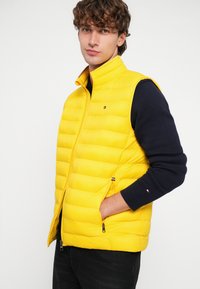 Gelbe Steppweste mit gesteppter Textur, Reißverschlusstaschen, Stehkragen und einem Logo-Detail. Wird über einem marineblauen Pullover getragen.