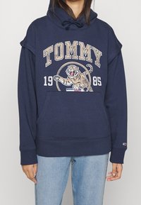 Sudadera con capucha de color azul marino que presenta una gran impresión gráfica de un tigre, el texto "TOMMY" y un bolsillo frontal tipo canguro. Tejido suave con puños acanalados.