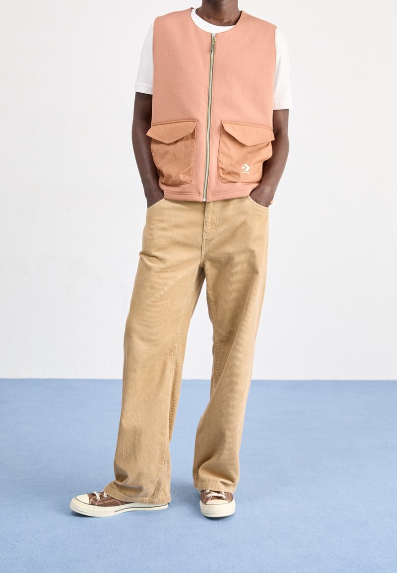 Homme portant une veste zippée pêche avec de grandes poches avant par-dessus une chemise blanche à manches courtes, un pantalon beige en velours côtelé et des baskets marron.