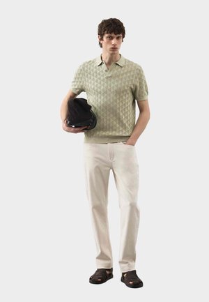 Giovane uomo con polo beige a fantasia, pantaloni chiari, sandali neri, tiene un cappello nero, una mano in tasca, in piedi davanti a uno sfondo uniforme.